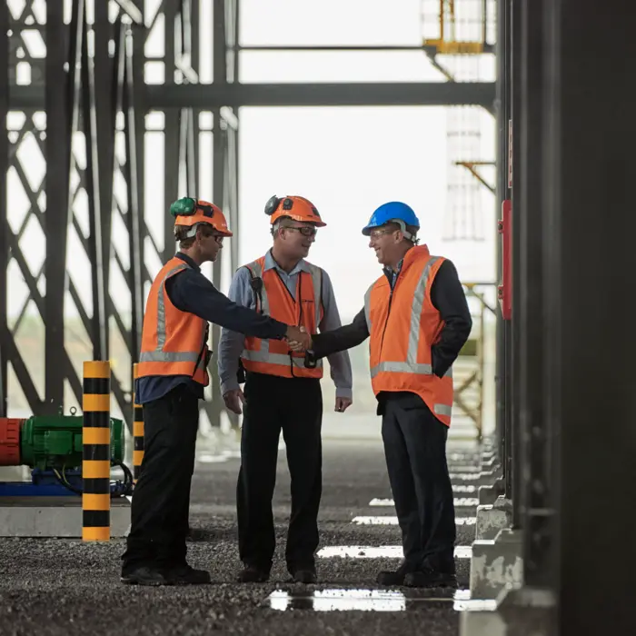 Three men in orange vests shaking hands at a construction site, symbolizing collaboration and agreement.
