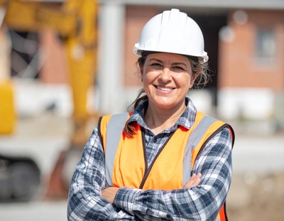 A woman wearing an orange vest and hard hat stands in front of a construction site, overseeing the ongoing work.
